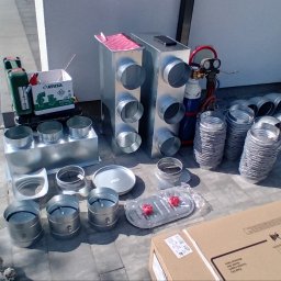 Bru-Nat Kamil Krawczyk - Przygotowane elementy wentylacyjne z blachy ocynkowanej, kształtki, rury spiro, skrzynki rozprężne, narzędzia i materiały montażowe rozłożone na szarych płytach chodnikowych, instalator rozpakowuje...