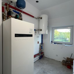 Bru-Nat Kamil Krawczyk - Biała pompa ciepła Panasonic Aquarea w nowym pomieszczeniu z instalacją hydrauliczną, zbiornikiem i oknem. Widoczne rury miedziane i PCV. Szare płytki podłogowe.