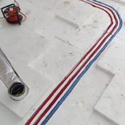 Bru-Nat Kamil Krawczyk - Ogrzewanie podłogowe: rury czerwone i niebieskie ułożone na białym styropianie, widoczny fragment wentylacji i zwinięty kabel na podłodze w trakcie instalacji.