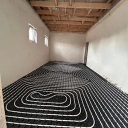 Białe rury ogrzewania podłogowego ułożone na czarnej macie w surowym wnętrzu z drewnianą konstrukcją stropu i oknami. Widok na instalację przed zalaniem posadzką.