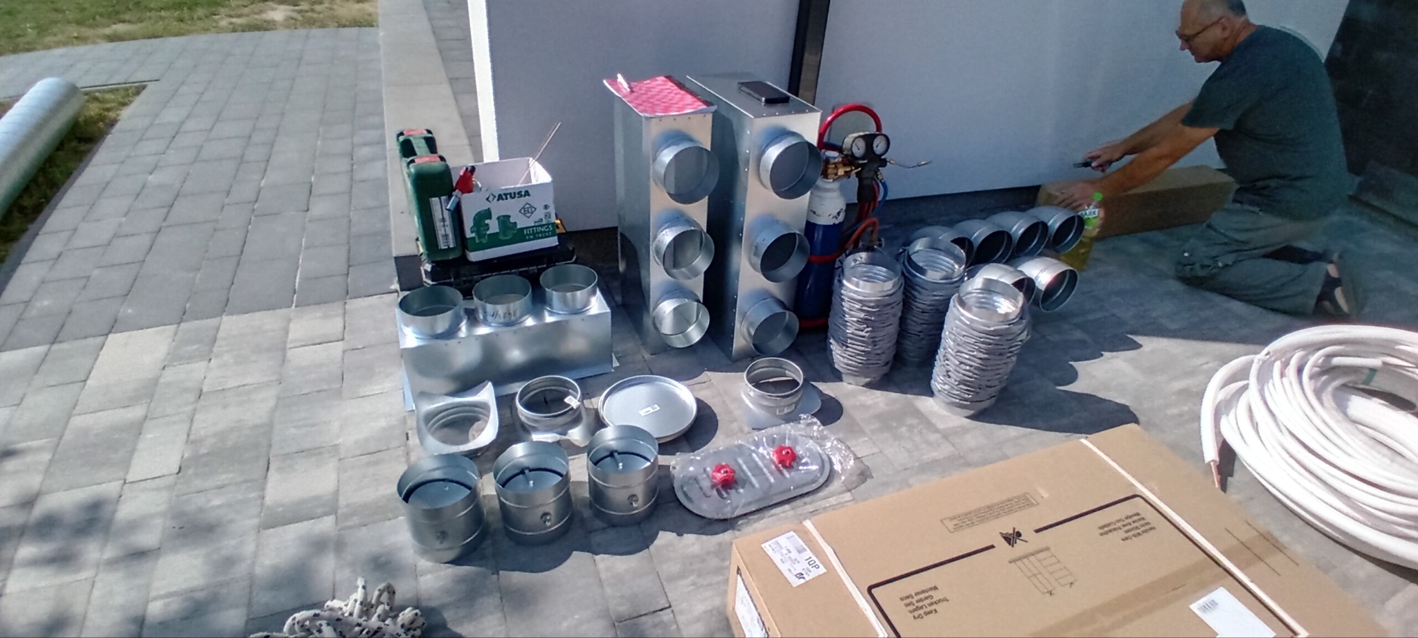 Przygotowane elementy wentylacyjne z blachy ocynkowanej, kształtki, rury spiro, skrzynki rozprężne, narzędzia i materiały montażowe rozłożone na szarych płytach chodnikowych, instalator rozpakowuje...