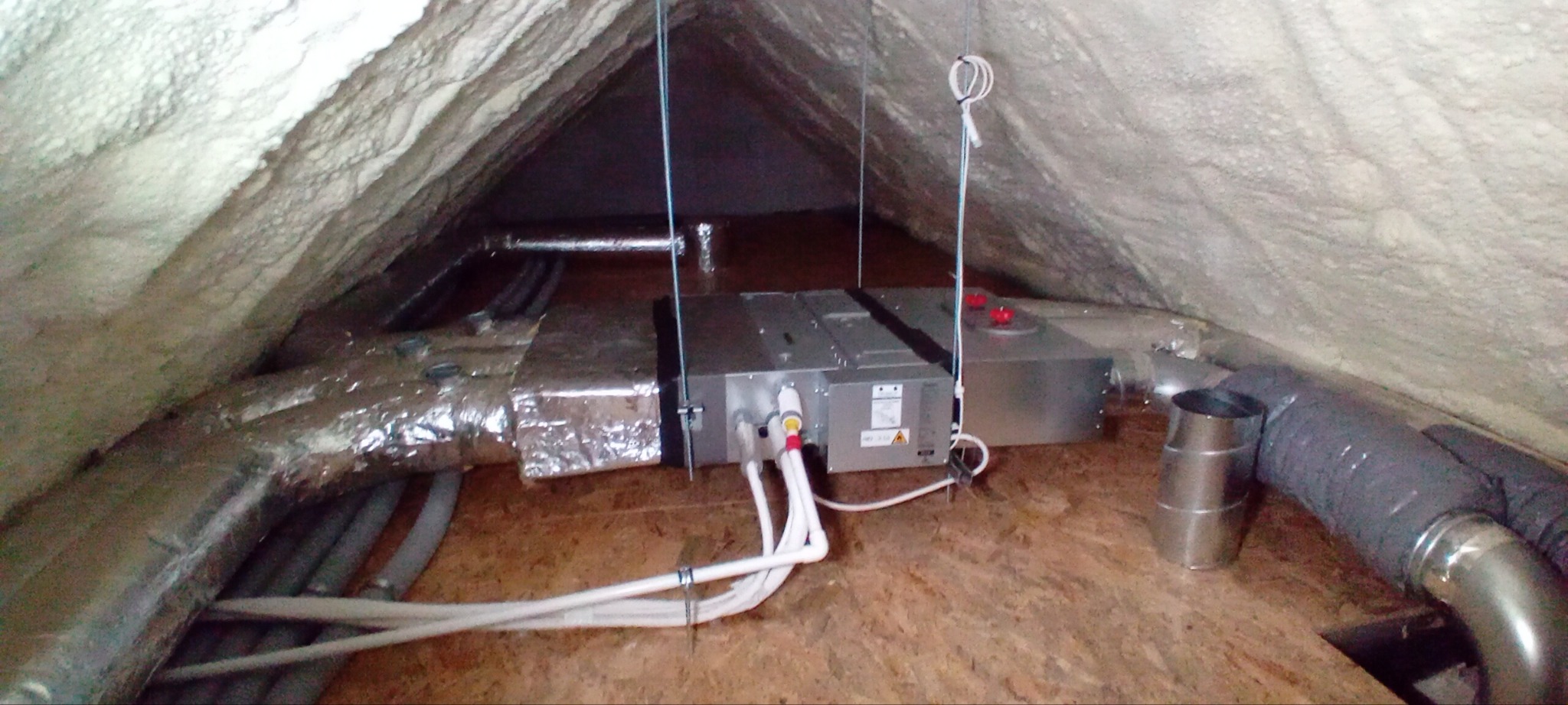 Instalacja w przestrzeni poddasza z widocznymi przewodami wentylacyjnymi, rurami oraz centralną jednostką z białymi przewodami. Ściany i dach pokryte pianką izolacyjną.