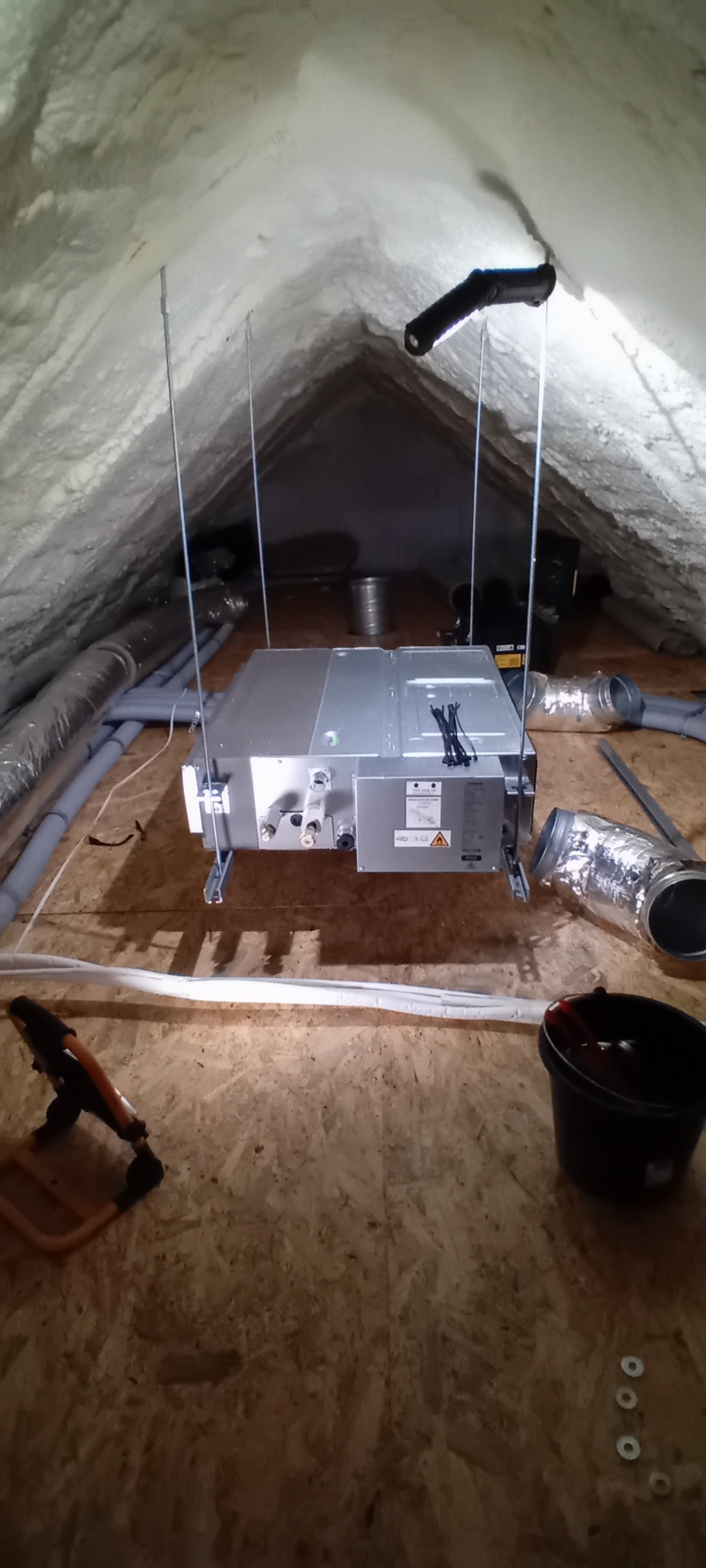 Montaż centrali wentylacyjnej z rekuperacją w przestrzeni poddasza ocieplonego pianą, widoczne elementy mocujące, kanały wentylacyjne i narzędzia pracy.