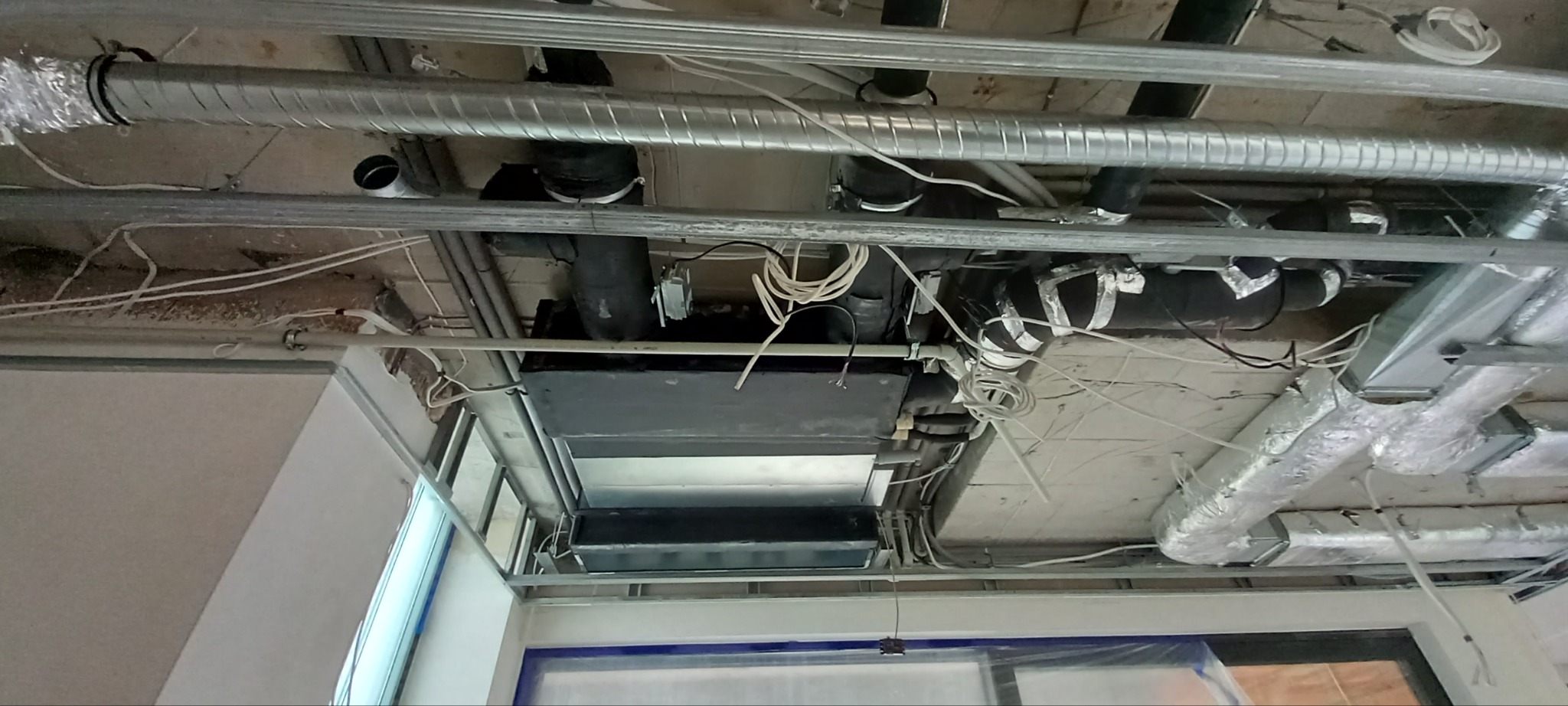 Widok sufitu w trakcie montażu instalacji wentylacyjnej z widocznymi przewodami wentylacyjnymi z folii aluminiowej, czarnymi rurami, przewodami elektrycznymi oraz elementami konstrukcji...