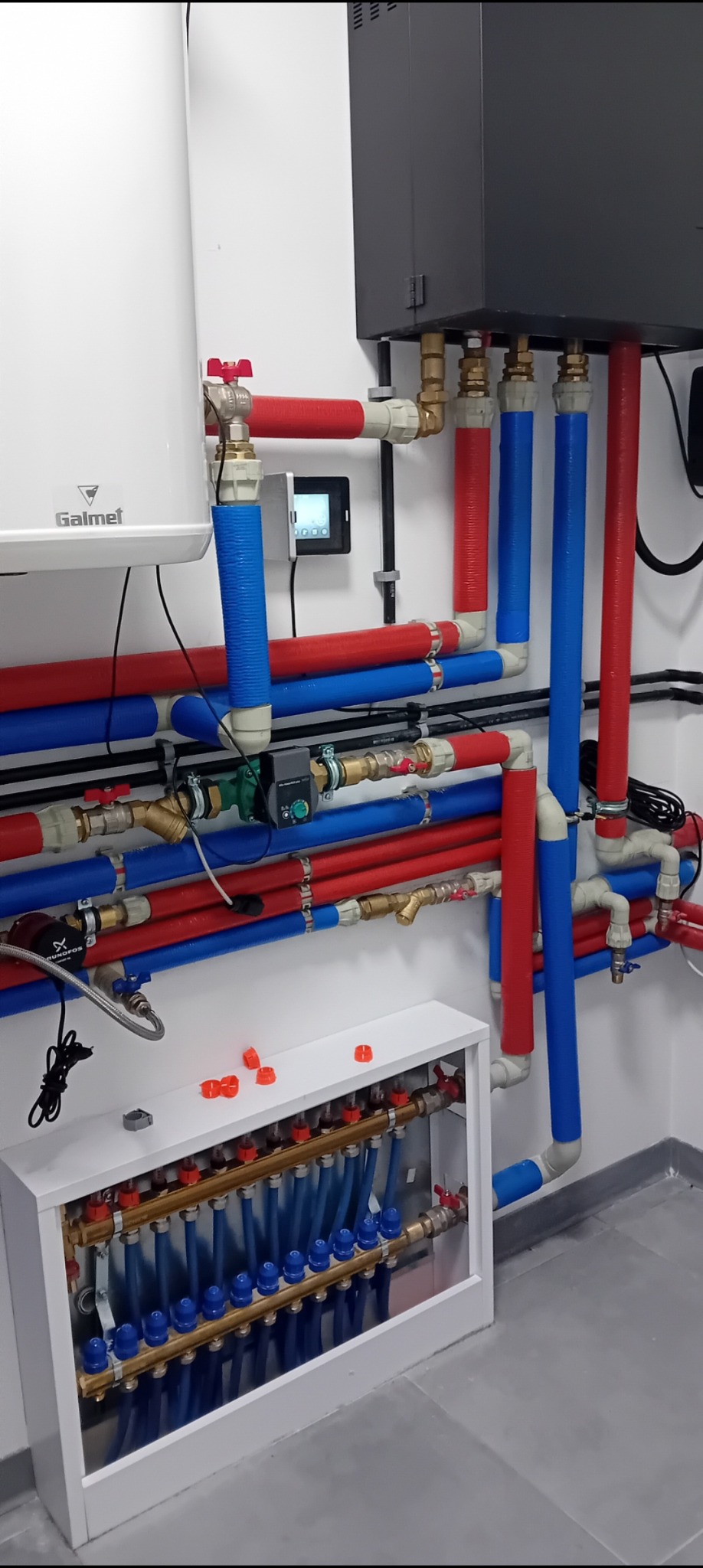 Rozdzielacz ogrzewania podłogowego w białej szafce, z widocznymi rurami w izolacji czerwonej i niebieskiej, oraz biały zasobnik ciepłej wody użytkowej Galmet na ścianie.