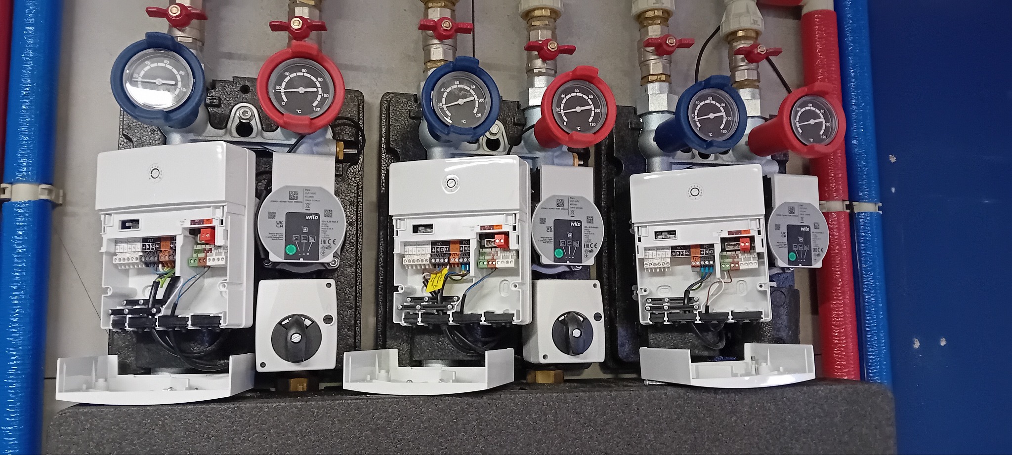 Rozdzielacz ogrzewania podłogowego z widocznymi manometrami i modułami sterującymi, zamontowany na szarej ścianie, z izolowanymi rurami w kolorze czerwonym i niebieskim.