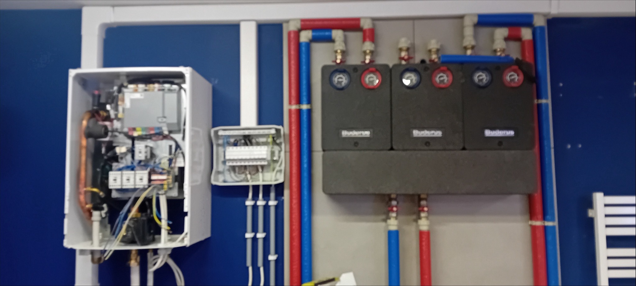 Otwarta obudowa kotła grzewczego z widocznymi elementami wewnętrznymi, obok skrzynka elektryczna i trzy moduły z logo Buderus, rury w kolorach czerwonym i niebieskim.