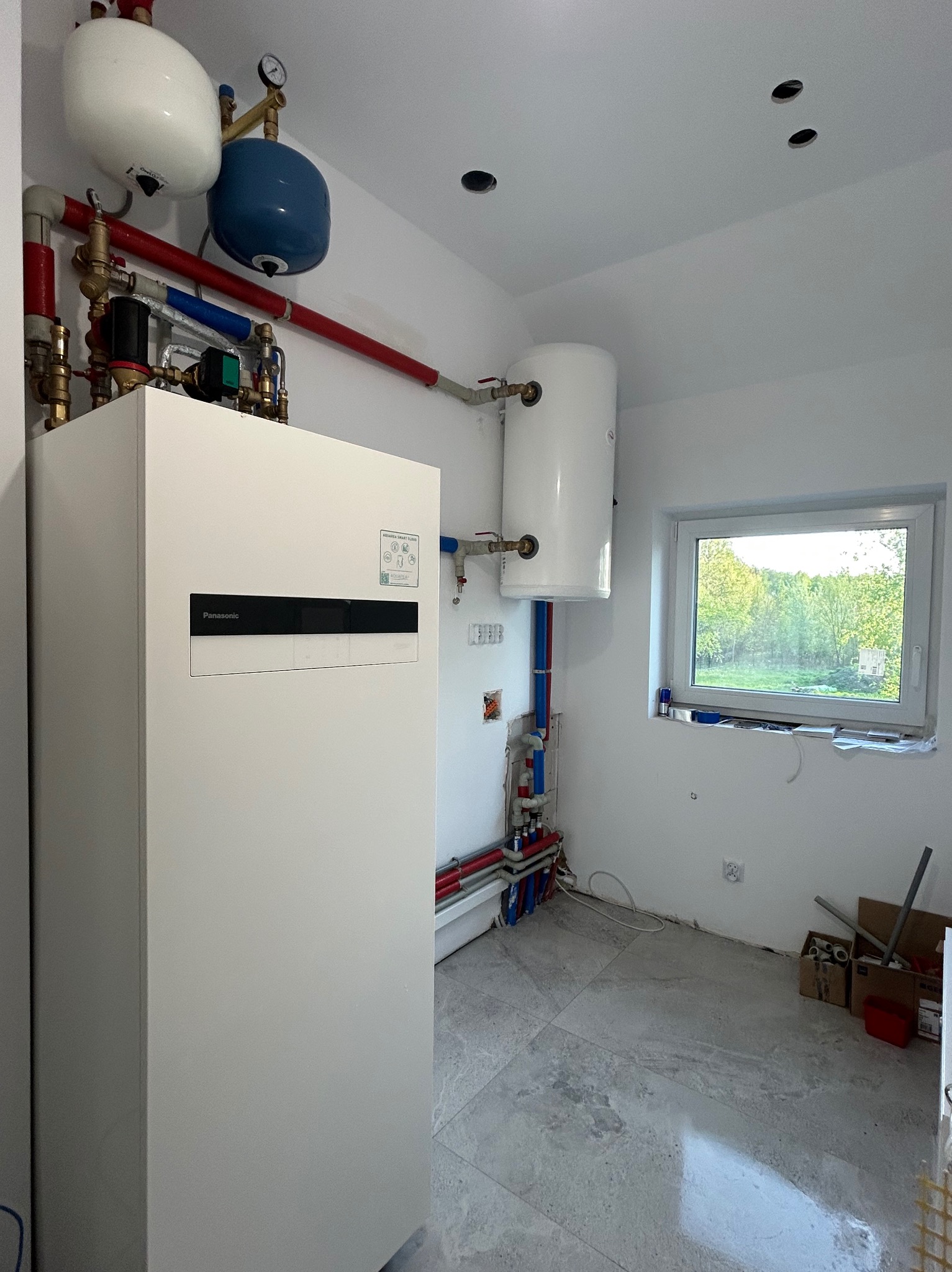 Biała pompa ciepła Panasonic Aquarea w nowym pomieszczeniu z instalacją hydrauliczną, zbiornikiem i oknem. Widoczne rury miedziane i PCV. Szare płytki podłogowe.