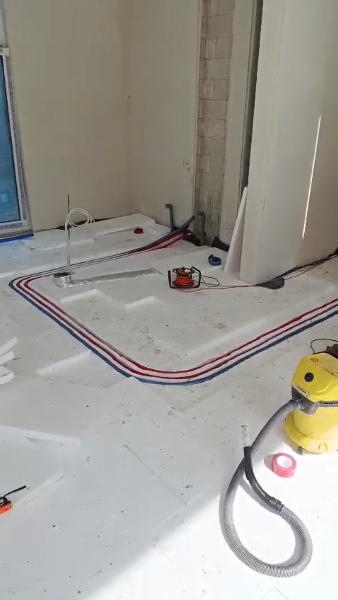 Układanie rur ogrzewania podłogowego na białym styropianie, widoczny odkurzacz i narzędzia. Instalacja w trakcie realizacji, przed wylaniem posadzki.