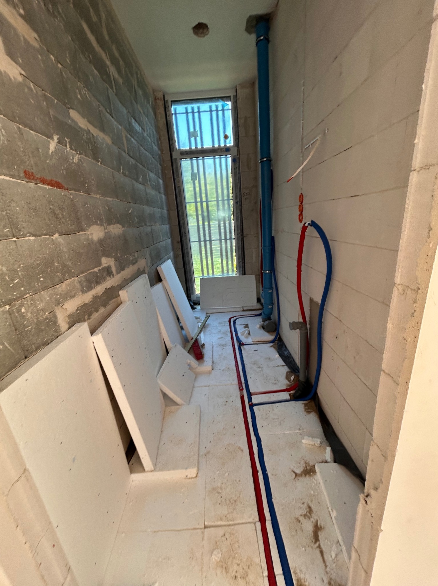 Wnętrze w trakcie prac: ułożone płyty izolacyjne pod ogrzewanie podłogowe, widoczne rury instalacji grzewczej, surowe ściany z betonu komórkowego i okno.