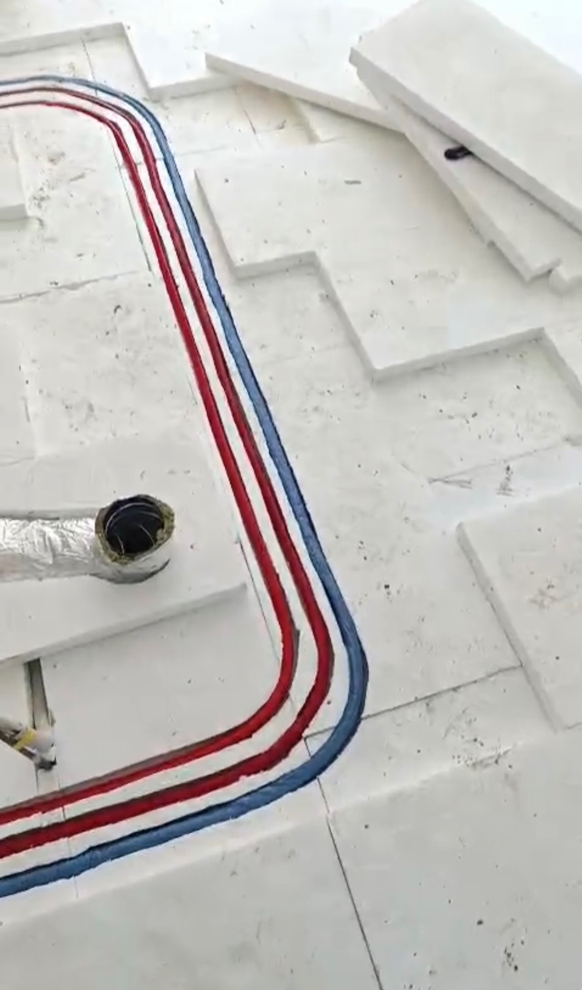 Ułożone rury ogrzewania podłogowego na białym styropianie, z widocznym otworem na rurę. Czerwone i niebieskie rury ułożone równolegle.
