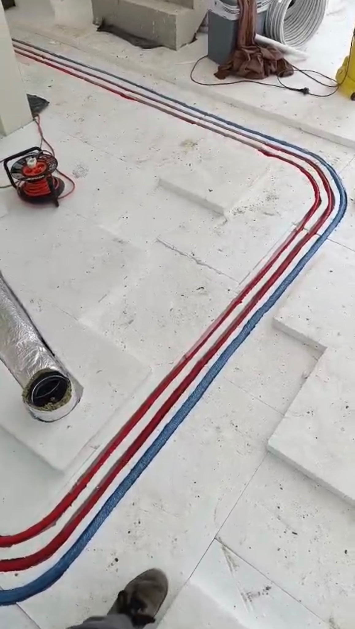 Ogrzewanie podłogowe: rury czerwone i niebieskie ułożone na białym styropianie, widoczny fragment wentylacji i zwinięty kabel na podłodze w trakcie instalacji.