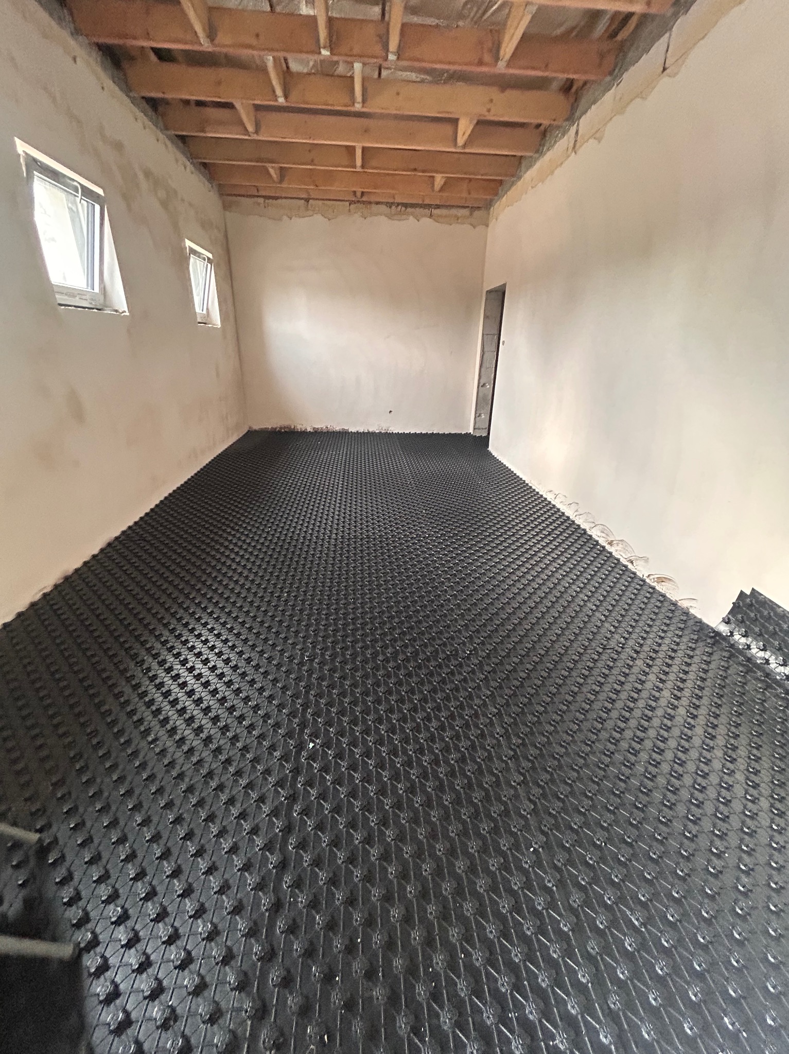 Czarna mata do ogrzewania podłogowego rozłożona w surowym pomieszczeniu z drewnianym stropem i dwoma małymi oknami. Widok z góry ukazuje regularny wzór maty.