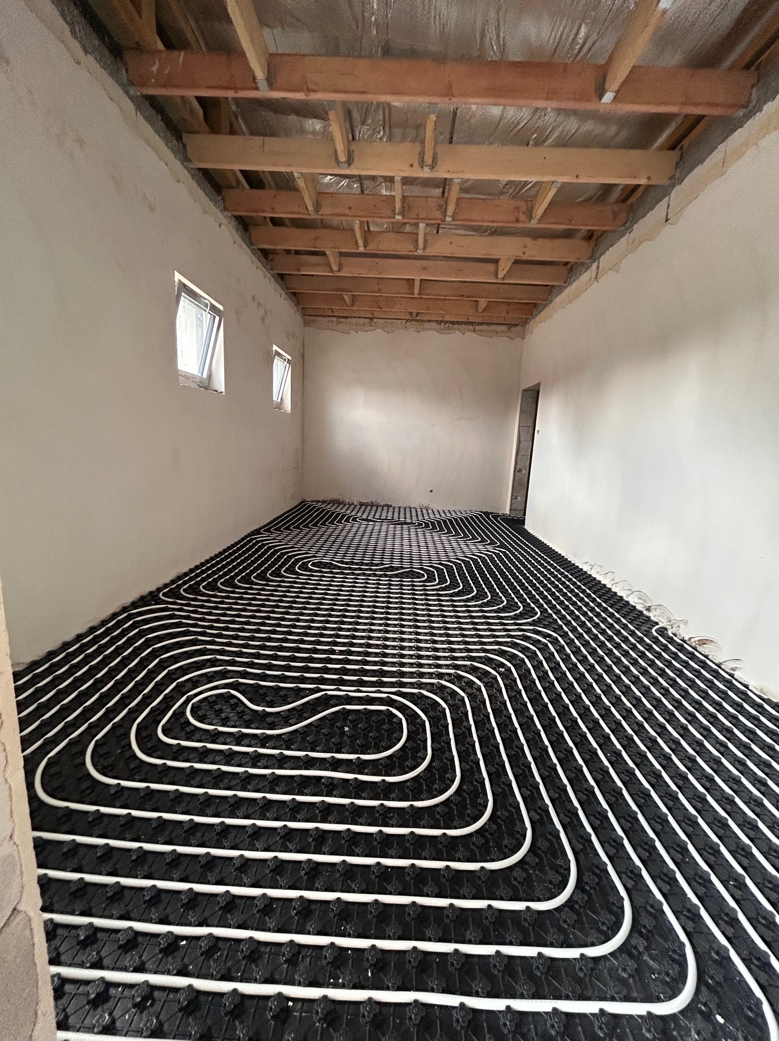 Białe rury ogrzewania podłogowego ułożone na czarnej macie w surowym wnętrzu z drewnianą konstrukcją stropu i oknami. Widok na instalację przed zalaniem posadzką.