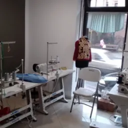 Wnętrze pracowni krawieckiej z dwoma maszynami do szycia, manekinem z ubraniem w tle i widokiem na ulicę przez okno.