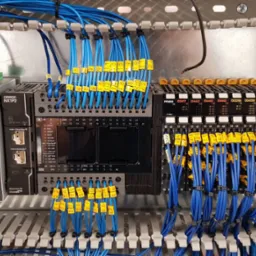 Szara szafa sterownicza z zamontowanymi modułami sterującymi Omron, niebieskimi kablami oznaczonymi żółtymi numerowanymi etykietami i listwami montażowymi