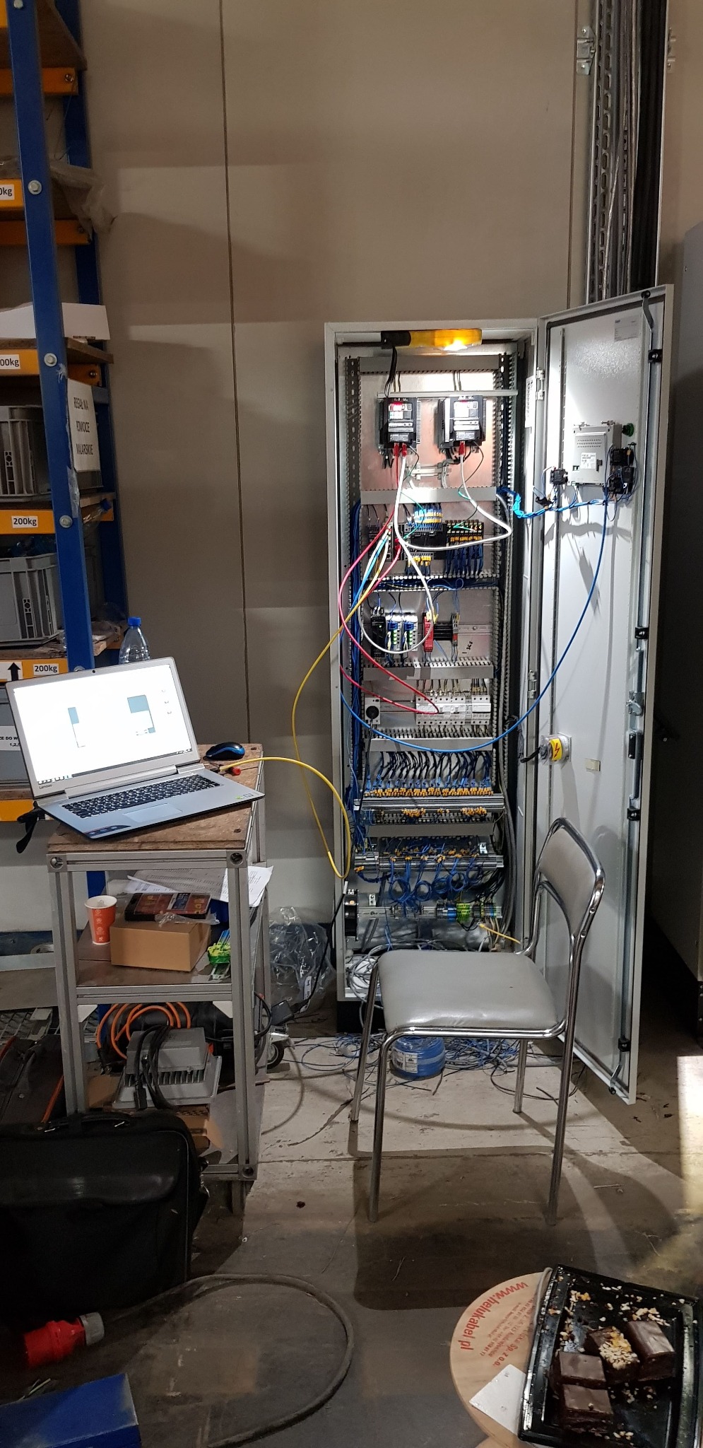 Otwarta szafa sterownicza z widocznymi elementami elektrycznymi, kablami i podłączeniami, obok laptop na wózku serwisowym i krzesło