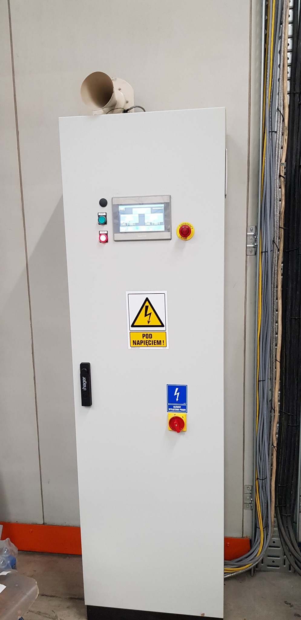 Biała szafa sterownicza z panelem dotykowym, przyciskami start/stop i awaryjnego wyłączenia, oraz naklejkami ostrzegawczymi 'Pod napięciem' i 'Główny wyłącznik prądu', z widoczną wiązką przewodów...