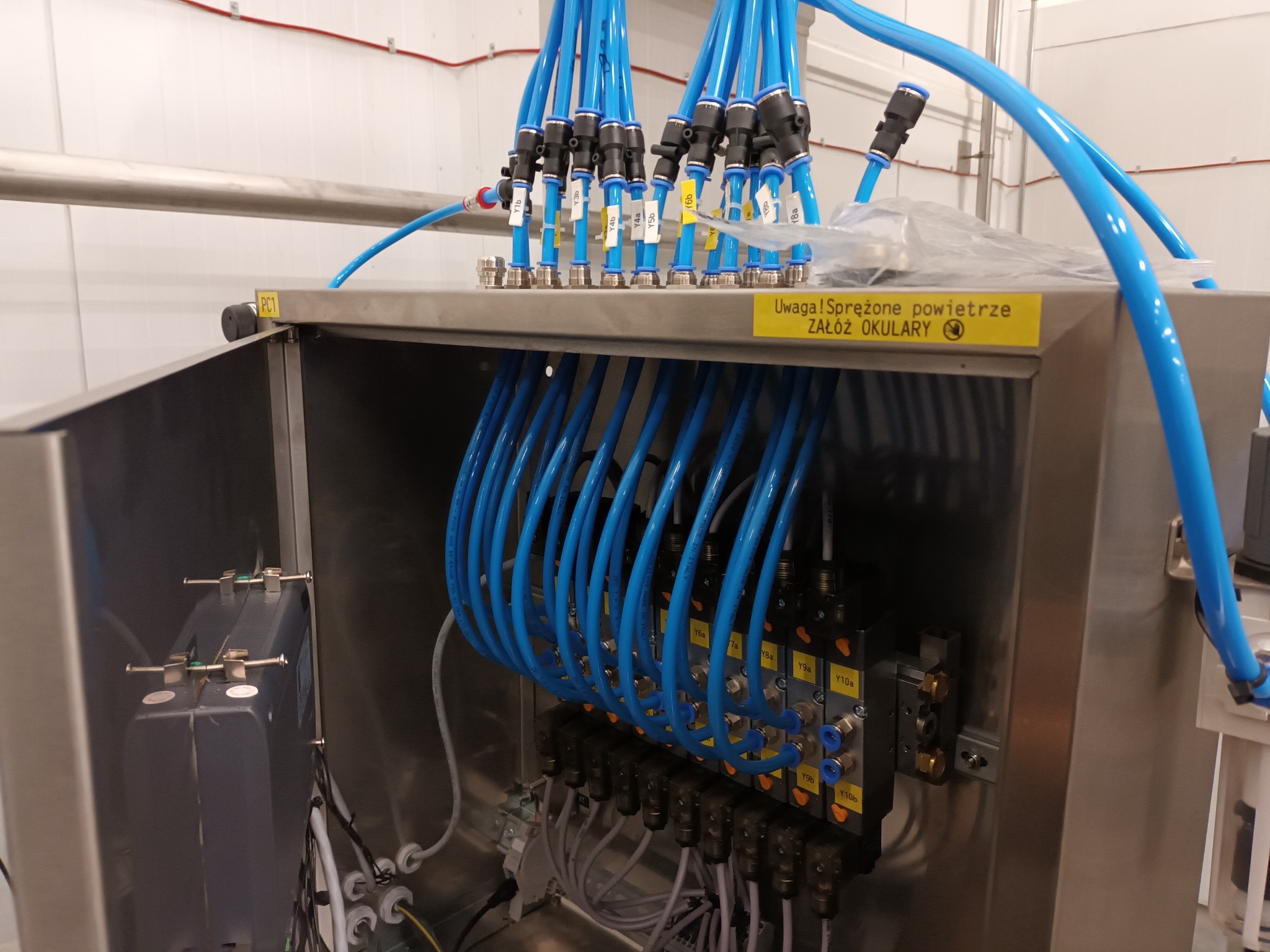 Wnętrze szafy sterowniczej z licznymi niebieskimi przewodami pneumatycznymi, zaworami elektromagnetycznymi z żółtymi etykietami i ostrzeżeniem o sprężonym powietrzu. Widoczna instalacja elektryczna...