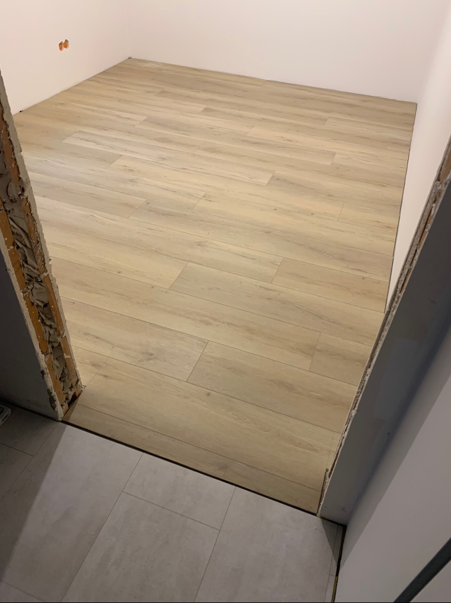 Jasne panele podłogowe ułożone w nowym pomieszczeniu, widoczne z perspektywy progu z surowym wykończeniem.