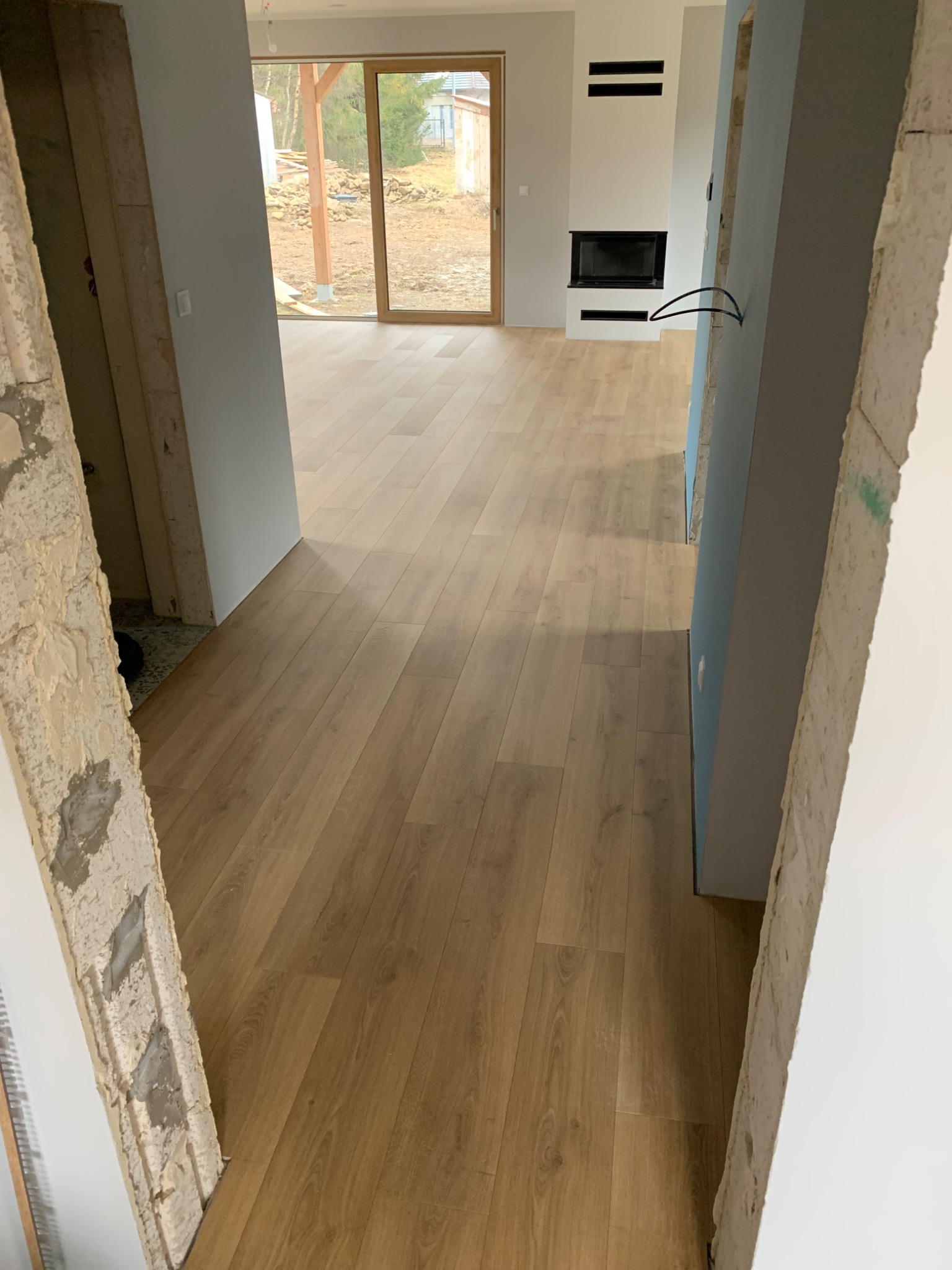 Jasne panele podłogowe ułożone w nowym domu, widoczne przez surowe otwory drzwiowe, z kominkiem w tle i widokiem na ogród przez duże okno.