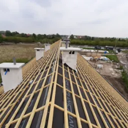 Widok z góry na konstrukcję dachu z drewnianych łat i czarną membraną, kominy z białego pustaka, w tle zabudowa jednorodzinna i samochody.