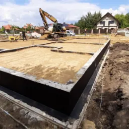 Fundamenty domu jednorodzinnego z hydroizolacją, przygotowane do dalszych prac budowlanych; w tle koparka i ubijarka gruntu.