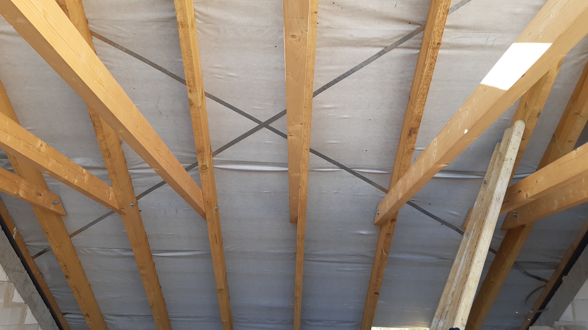 Konstrukcja dachu z belek drewnianych, zabezpieczona folią, widok od wewnątrz z fragmentem drabiny opartą o jedną z belek.
