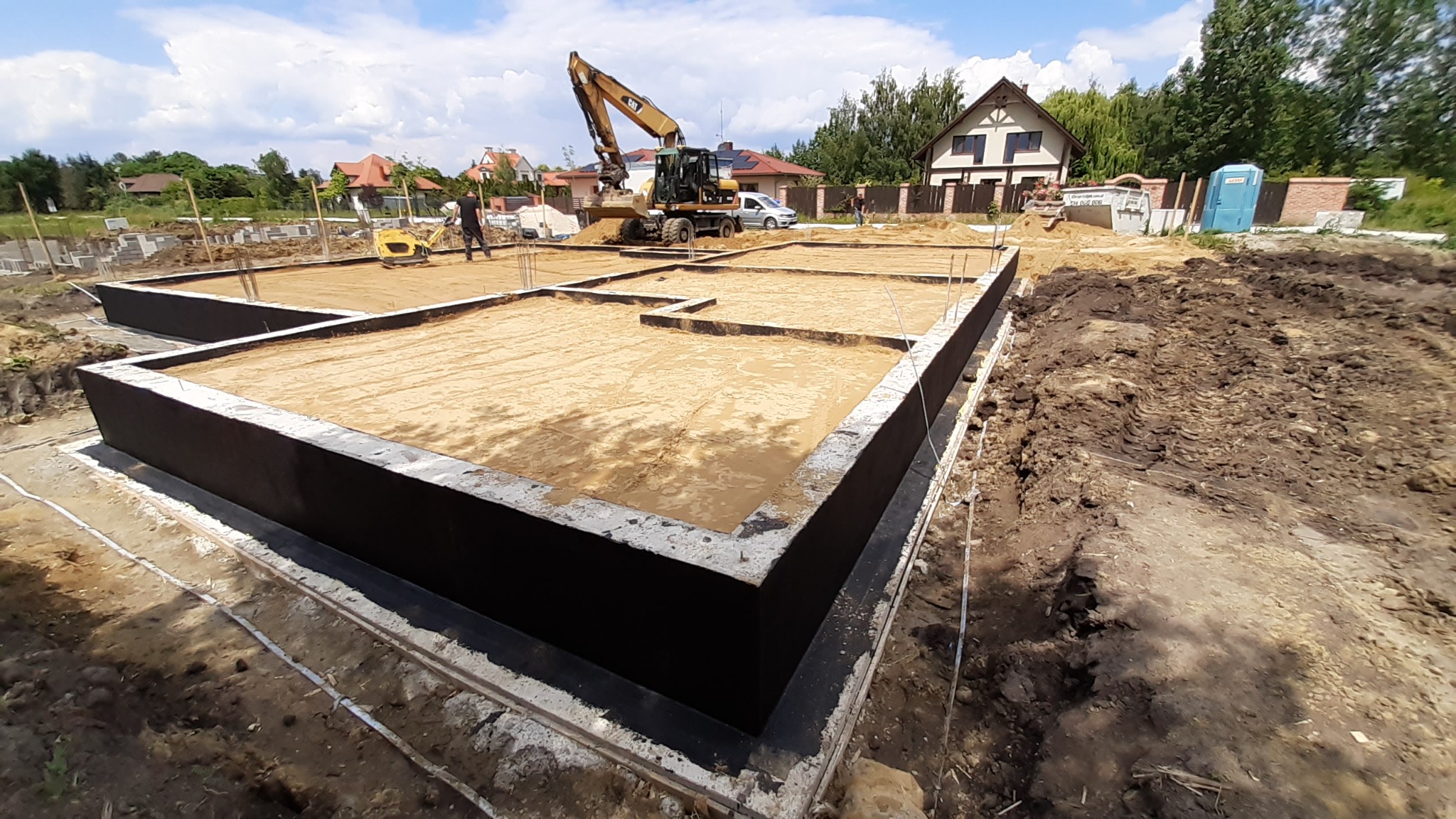 Fundamenty domu jednorodzinnego z hydroizolacją, przygotowane do dalszych prac budowlanych; w tle koparka i ubijarka gruntu.