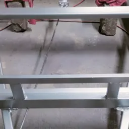 Metalowa konstrukcja pomalowana na szaro, wsparta na kamieniach z czerwonymi tkaninami. Widoczne spawy i przewody. Detal z bliska, surowe otoczenie.