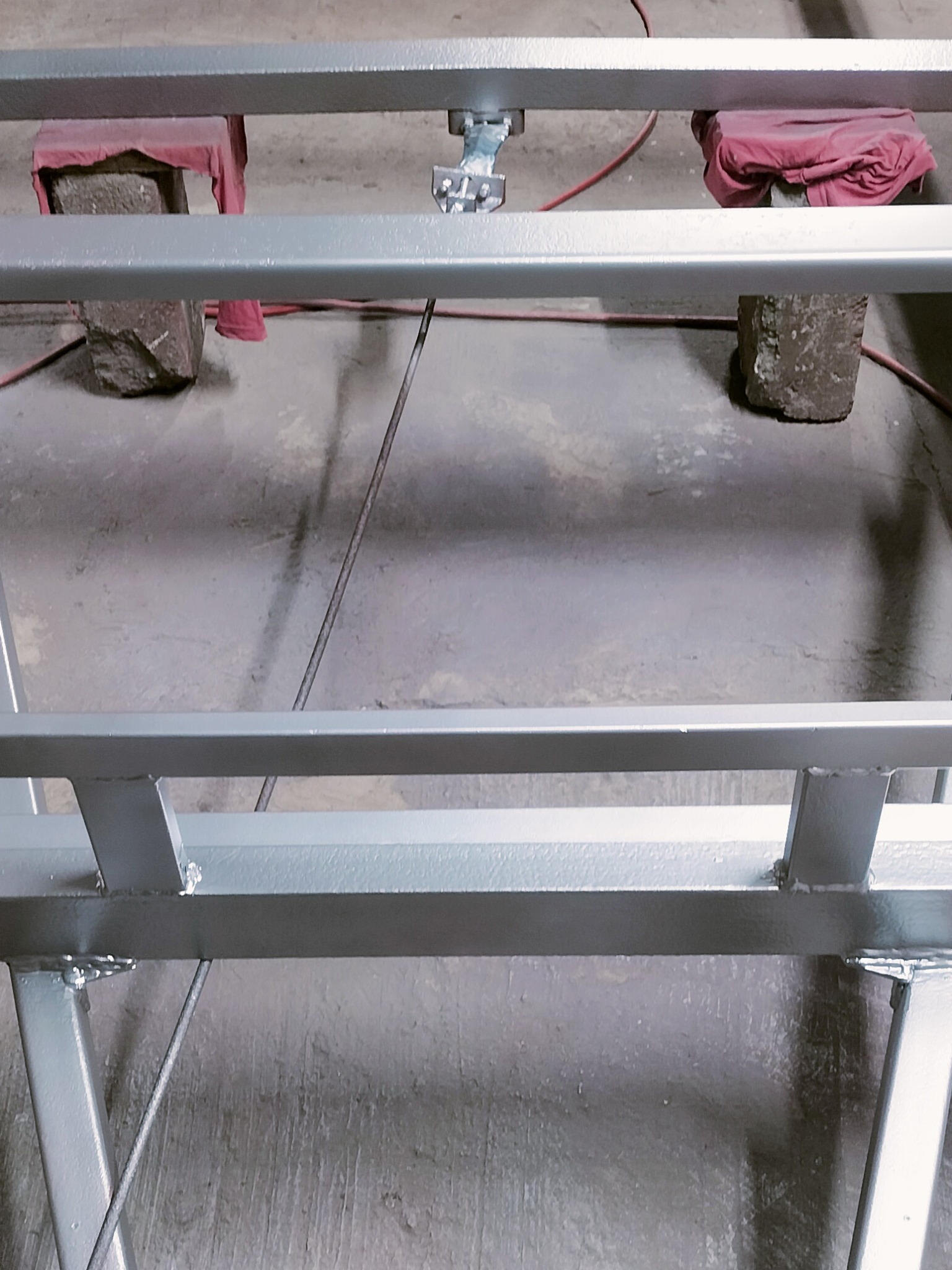 Metalowa konstrukcja pomalowana na szaro, wsparta na kamieniach z czerwonymi tkaninami. Widoczne spawy i przewody. Detal z bliska, surowe otoczenie.