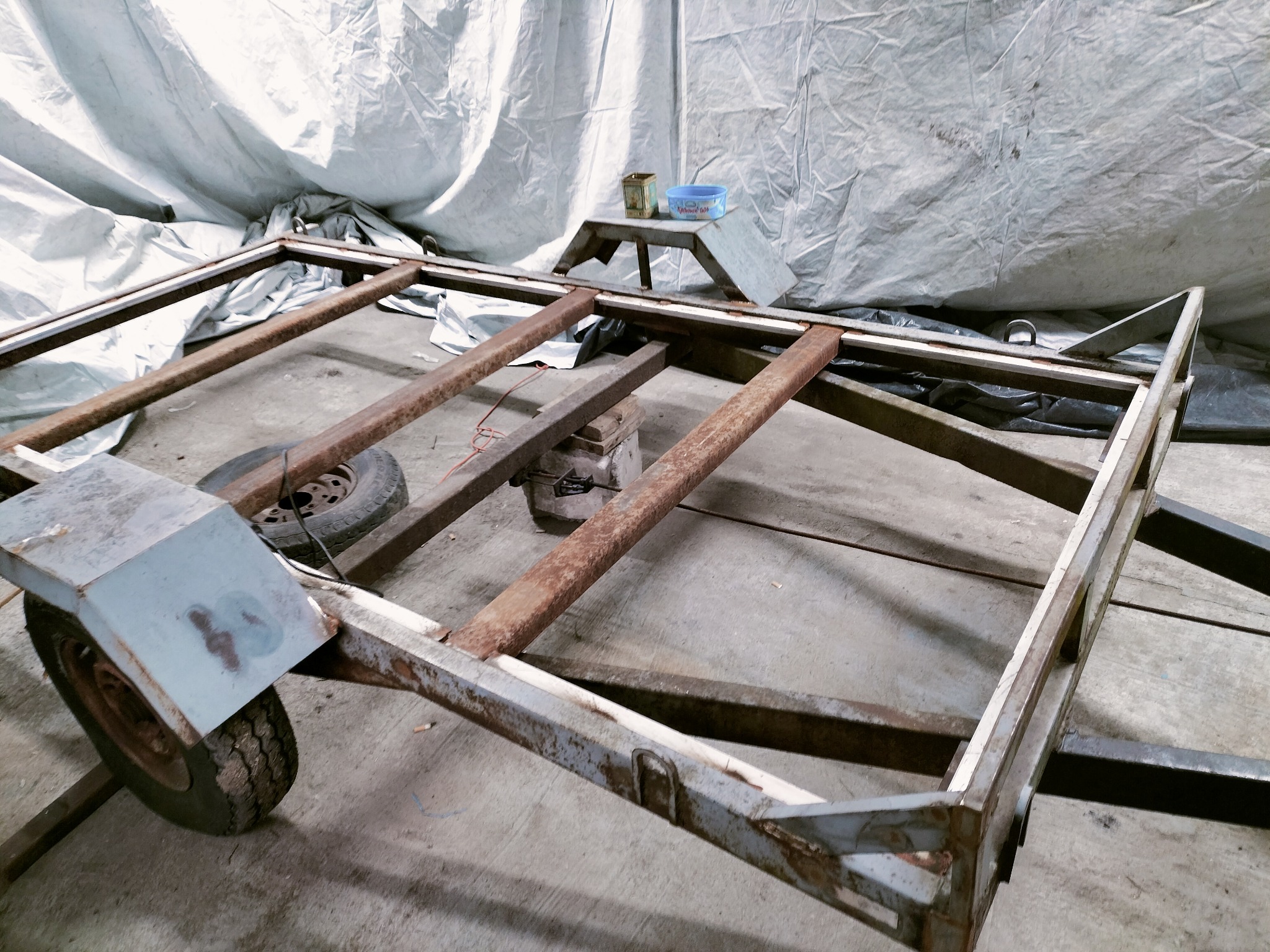 Rama przyczepy w warsztacie, widoczne ślady rdzy i wcześniejszej eksploatacji. Konstrukcja metalowa, częściowo pomalowana na szaro, z kołem po lewej stronie. W tle plandeka.