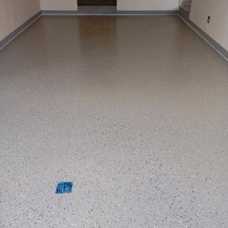 Osoba prywatna - Jasnoszara posadzka z drobnymi, ciemnymi wtrąceniami, wykonana w Nowym Dworze Mazowieckim, z widocznym niebieskim elementem na powierzchni i fragmentem ściany w tle.