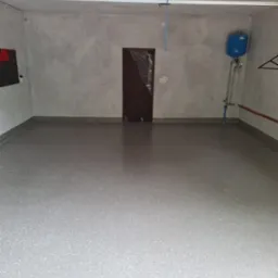 Jasnoszara, gładka podłoga w pustym pomieszczeniu, prawdopodobnie garażu, z widoczną instalacją hydrauliczną na ścianie i drzwiami zabezpieczonymi folią ochronną.