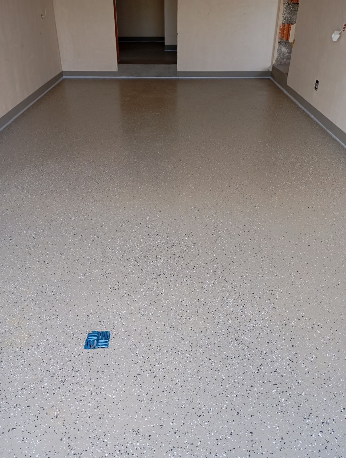 Jasnoszara posadzka z drobnymi, ciemnymi wtrąceniami, wykonana w Nowym Dworze Mazowieckim, z widocznym niebieskim elementem na powierzchni i fragmentem ściany w tle.