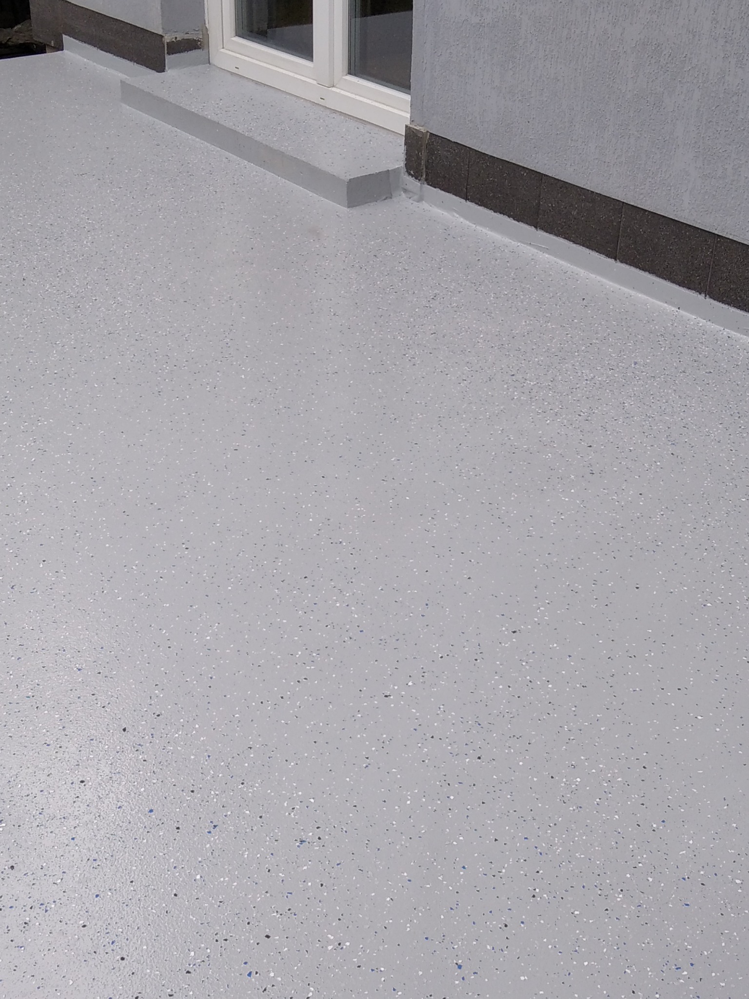 Jasnoszara, gładka powierzchnia z delikatnym, kolorowym wzorem, prawdopodobnie wykonana z żywicy, przylegająca do ściany budynku z oknem i niskim progiem.