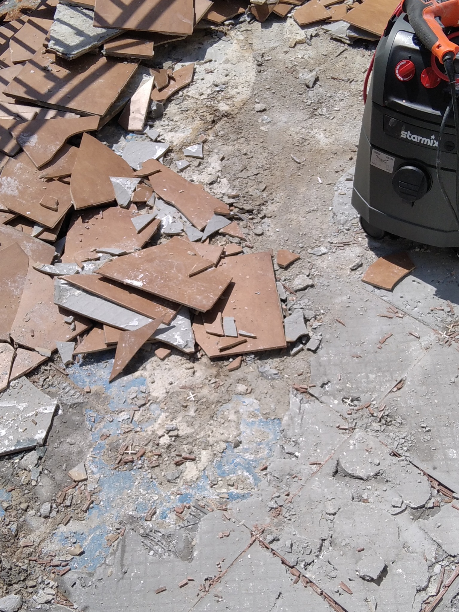 Demontaż starej posadzki z płytek ceramicznych, widoczne fragmenty płytek i resztki kleju, obok stoi odkurzacz przemysłowy Starmix.