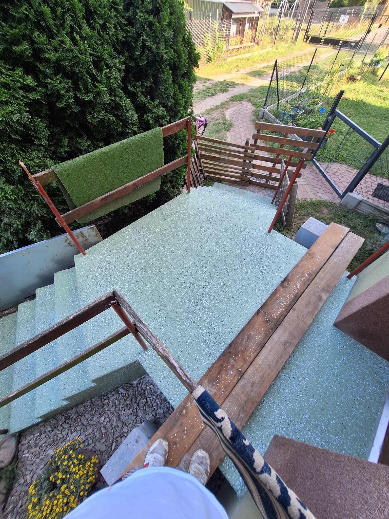 Jasnozielona powierzchnia schodów z żywicy, widok z góry, z drewnianymi deskami i wiaderkiem, w tle zieleń ogrodu.