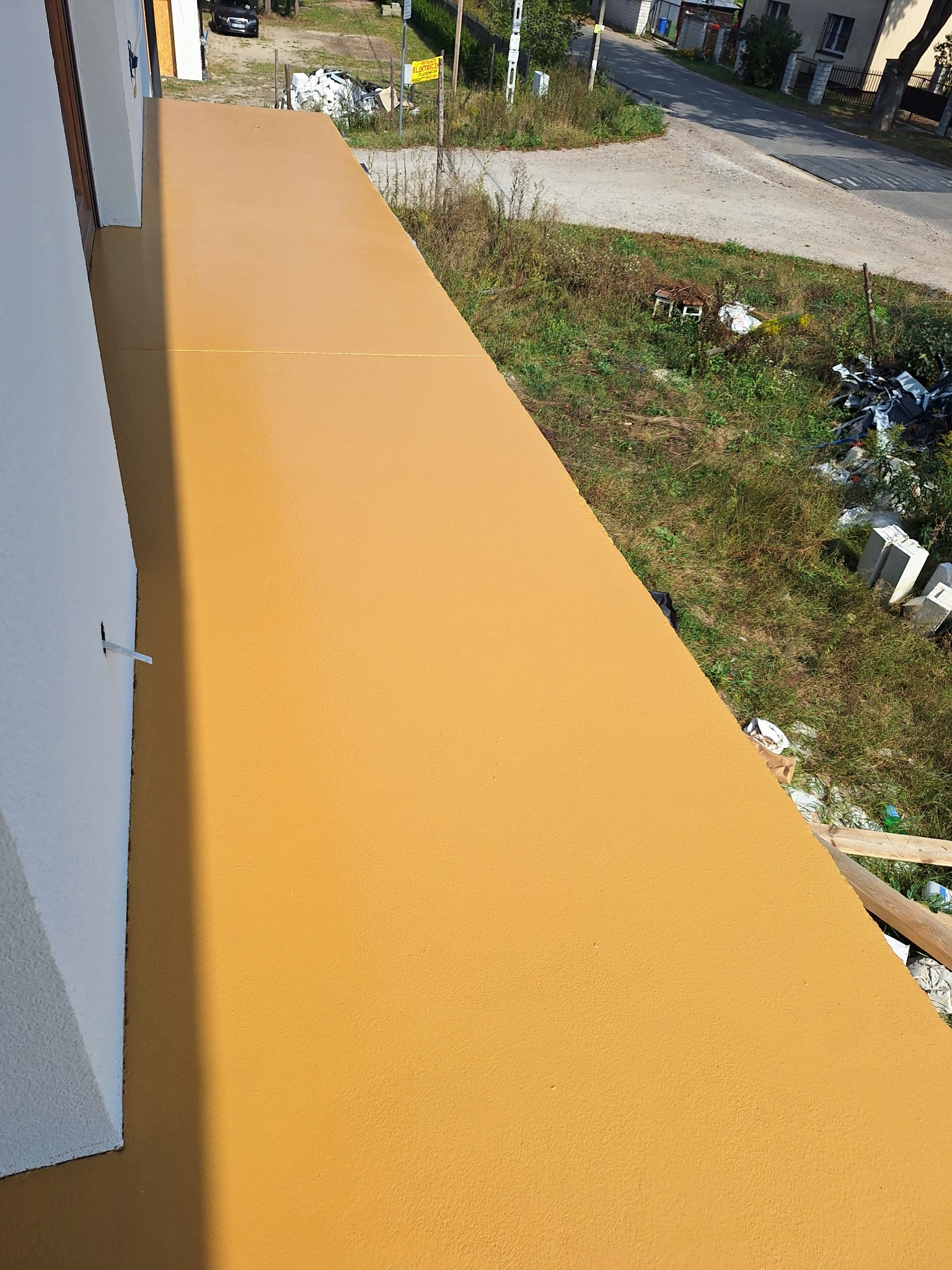 Widok z góry na balkon z jednolitą, pomarańczową powierzchnią. Widoczne połączenie z elewacją budynku i otoczenie z zielenią oraz elementami zabudowy mieszkalnej w tle.