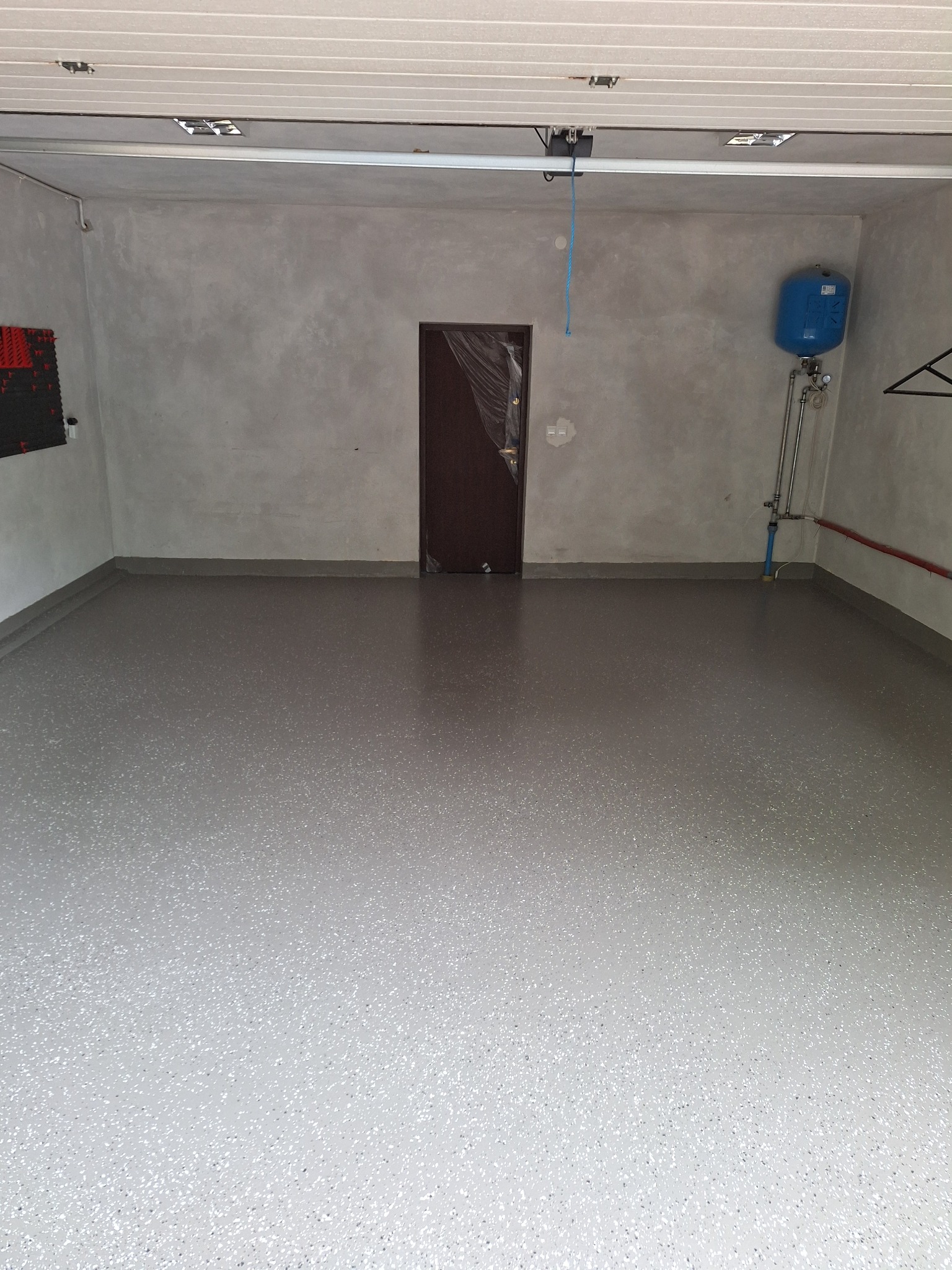 Jasnoszara, gładka podłoga w pustym pomieszczeniu, prawdopodobnie garażu, z widoczną instalacją hydrauliczną na ścianie i drzwiami zabezpieczonymi folią ochronną.