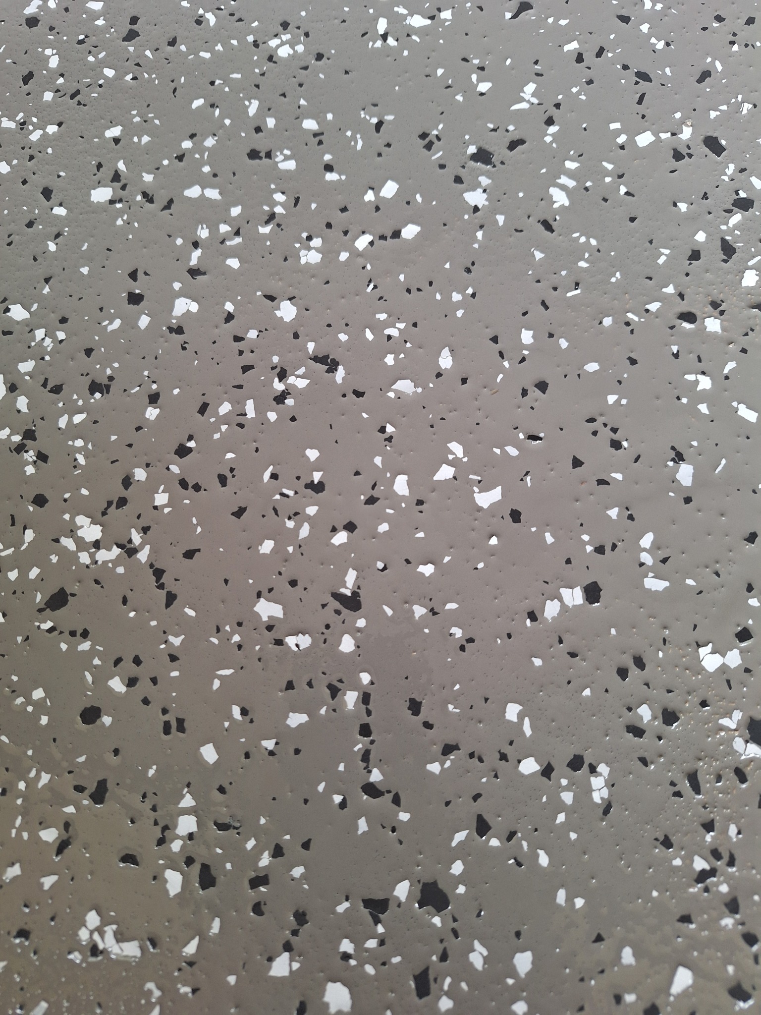 Gładka, szara posadzka z drobnymi, nieregularnymi wtrąceniami w kolorze białym i czarnym, tworzącymi efekt terrazzo.
