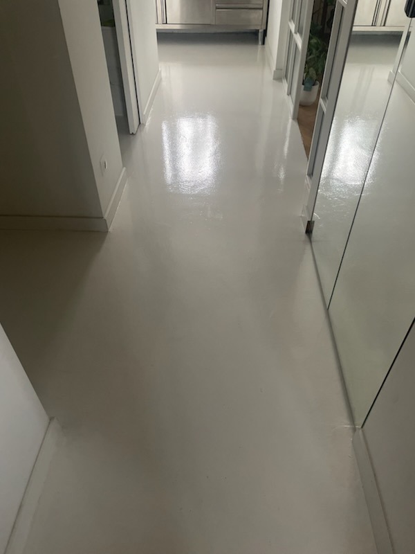 Jasnoszara, błyszcząca podłoga w korytarzu, z widocznym odbiciem światła i fragmentami drzwi oraz roślin w tle.
