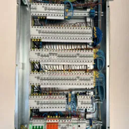 Otwarta skrzynka elektryczna w metalowej obudowie, zawierająca liczne bezpieczniki, kable w kolorach zielono-żółtym, brązowym i niebieskim, ułożone poziomo na kilku poziomach, zamontowana na ścianie.