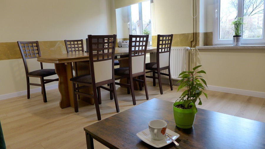 Jadalnia w Tychach z drewnianym stołem, krzesłami z kratką na oparciach i filiżanką na stoliku kawowym, w tle okno z roślinami.