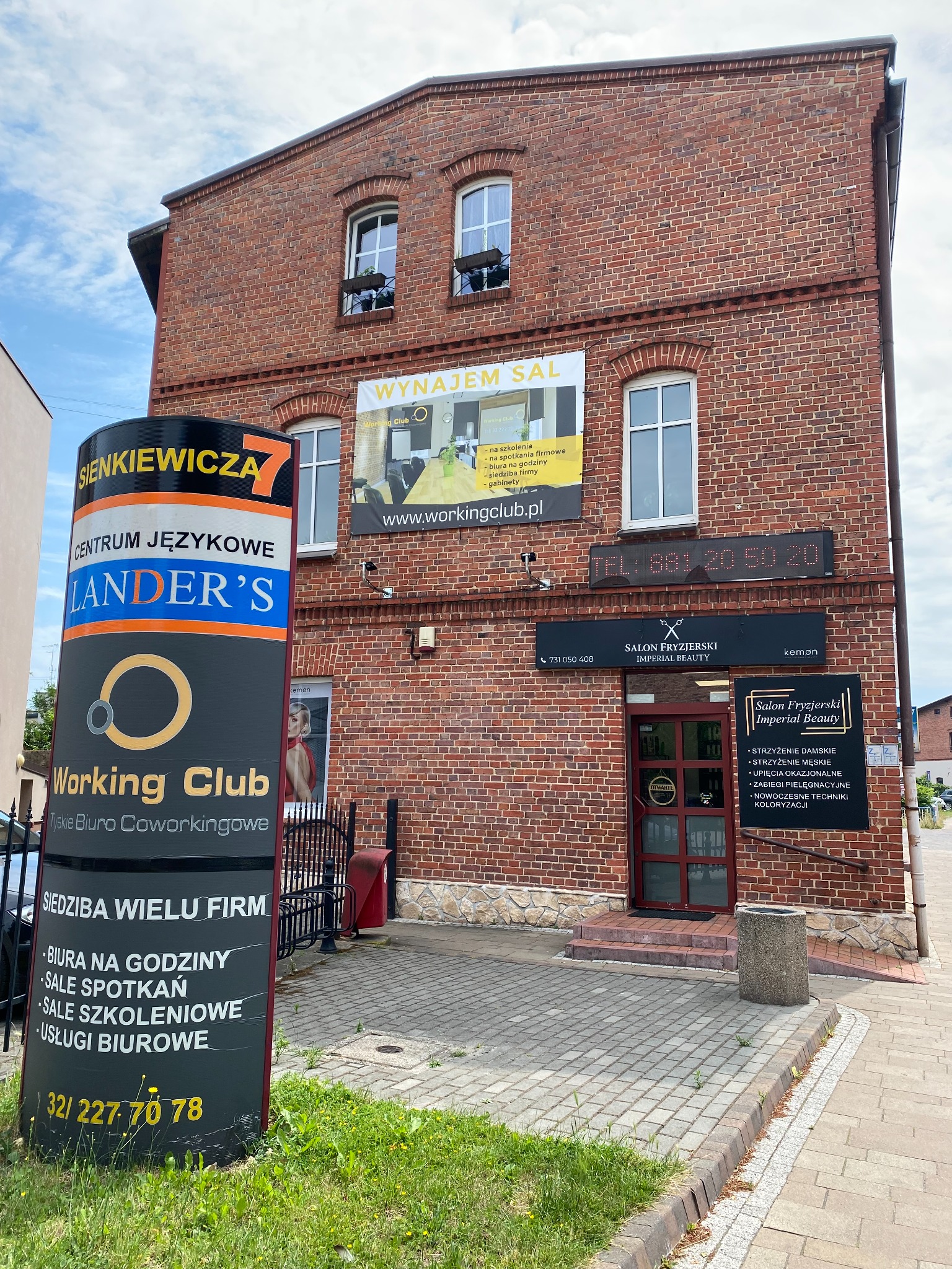 Zewnętrzny widok dwupiętrowego budynku z czerwonej cegły z szyldami firm: centrum językowe, biuro coworkingowe i salon fryzjerski. Na elewacji baner reklamujący wynajem sal. Przed budynkiem tablica...