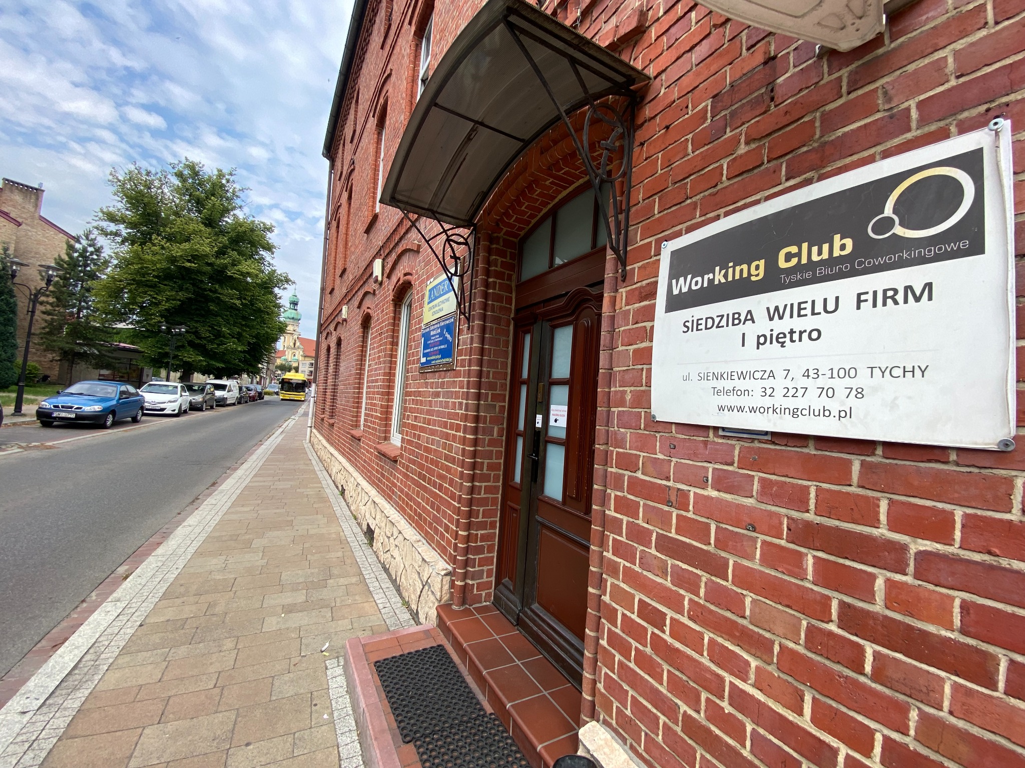 Budynek biurowy z czerwonej cegły z szyldem 'Working Club Tyskie Biuro Coworkingowe' oraz informacją o siedzibie wielu firm, zadaszenie nad wejściem.