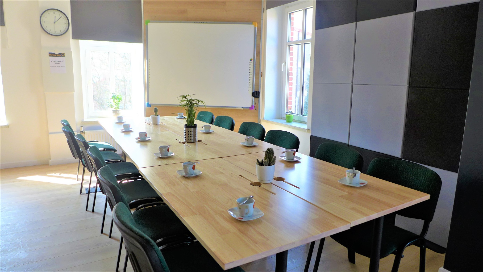 Jasna sala konferencyjna z dużym drewnianym stołem, zielonymi krzesłami, białą tablicą, oknami i dekoracyjnymi panelami akustycznymi na ścianie. Na stole ustawione filiżanki z kawą i rośliny...