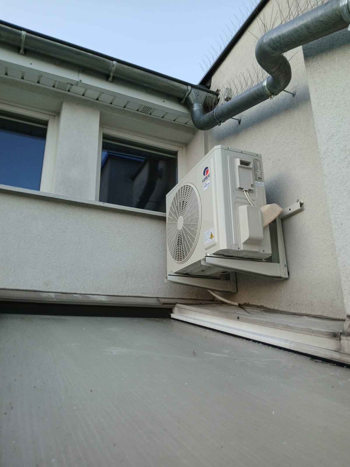 Zewnętrzna jednostka klimatyzacji marki Gree zamontowana na elewacji budynku, widoczna z perspektywy dachu, z rynną i zabezpieczeniami przed ptakami w tle.