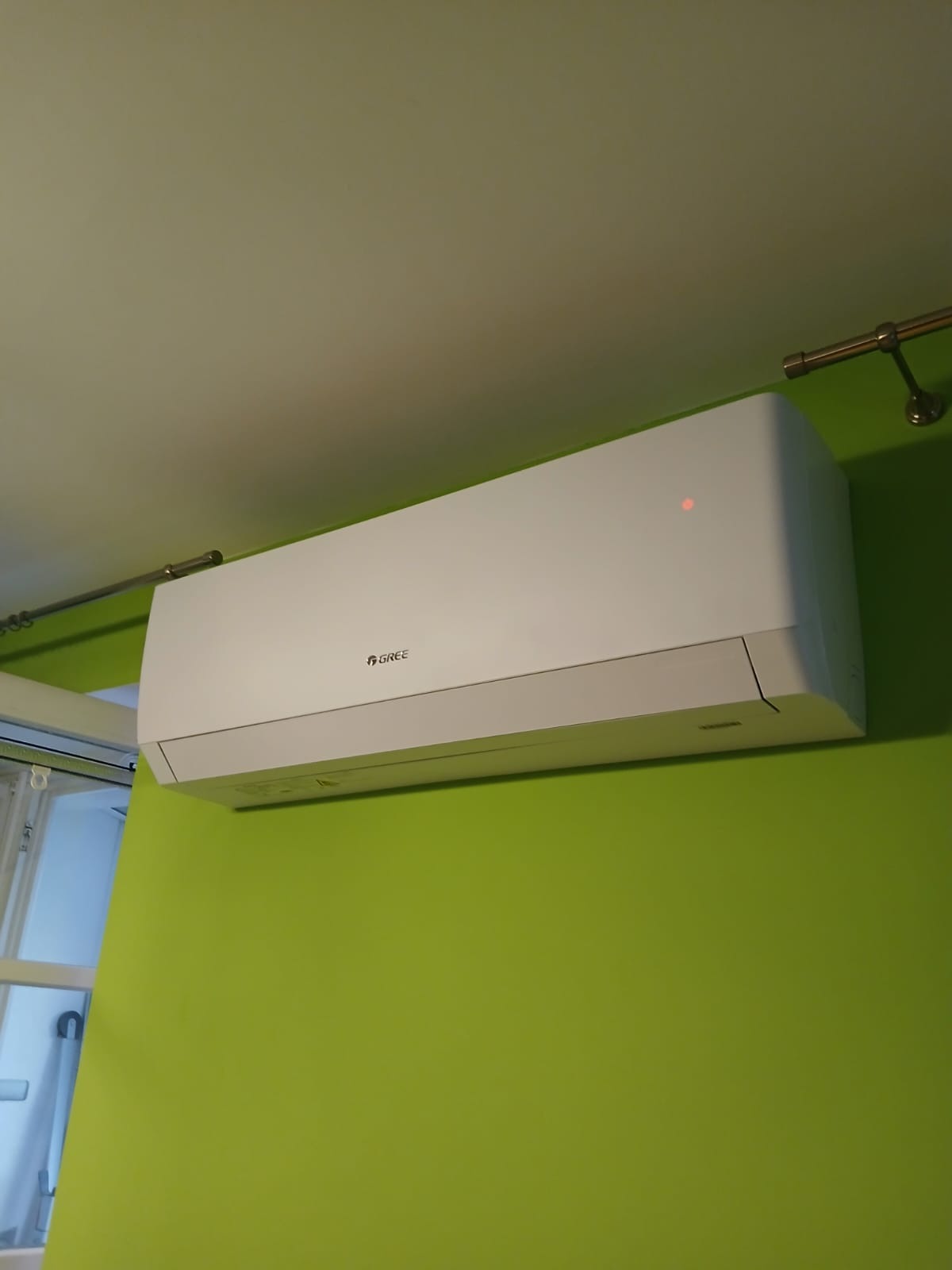 Biała jednostka wewnętrzna klimatyzacji marki Gree zamontowana na limonkowo-zielonej ścianie w pomieszczeniu.