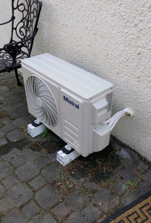 Jednostka zewnętrzna klimatyzacji Mistral zamontowana na betonowych wspornikach przy ścianie budynku w Warszawie, widoczne przewody i fragment dekoracyjnego krzesła.